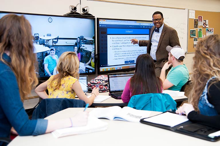 a professor teaches their students in front of a pair of large monitors.