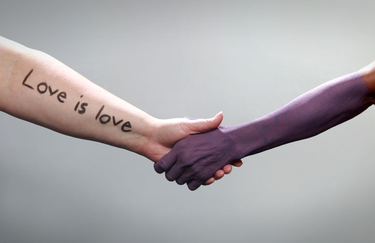 An arm with 'Love is love' written on it extends and is holding the hand of another extended arm painted purple.